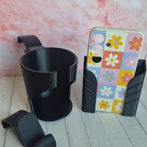以下が含まれることがあります： 黒色の3Dプリントされたカーアクセサリーセット。カップホルダー、電話ホルダー、クリップが含まれています。電話ホルダーには、カラフルな花柄のケースが付いた電話が入っています。アイテムは、ピンクの背景の白い木製の表面に表示されています。
