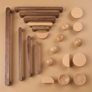 Può includere: Un assortimento di ferramenta per mobili in legno, tra cui maniglie, pomelli e maniglie a mezzaluna. Le maniglie variano in lunghezza, con la più lunga che misura circa 30 cm. I pomelli sono sferici e circolari e le tonalità del legno variano da chiare a scure.