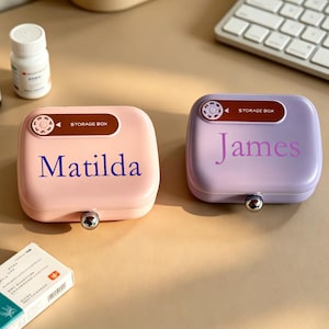 May include: Two personalized storage boxes, one pink with "Matilda" in blue, the other lavender with "James" in purple. Each box has a silver clasp and a "STORAGE BOX" label. A white pill bottle and a box of medication are also visible.