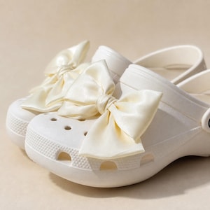 May include: A pair of off-white slip-on clogs featuring a smooth, matte finish and a series of circular ventilation holes along the front. Each clog is adorned with a large, lustrous, satin-like fabric bow in a matching off-white color.