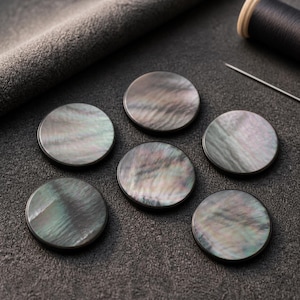 May include: Six circular buttons crafted from iridescent mother-of-pearl shell. Each button features a smooth, polished surface with a swirling, dark gray and charcoal color palette that displays subtle hints of green and purple light reflections.