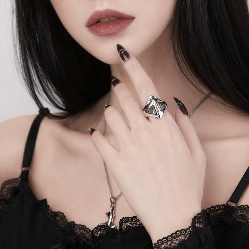 May include: A silver-toned ring featuring a geometric, multi-faceted, and angular design with open-work detailing. The ring is worn on a finger, accompanied by a silver-toned chain necklace with a pendant, and a black lace-trimmed garment.