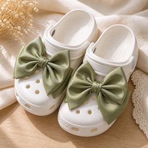 May include: A pair of white slip-on clogs featuring large, sage green satin bows attached to the top of each shoe. The clogs have a perforated toe box, a rounded silhouette, and a pivoting heel strap, showcasing a smooth, matte finish.