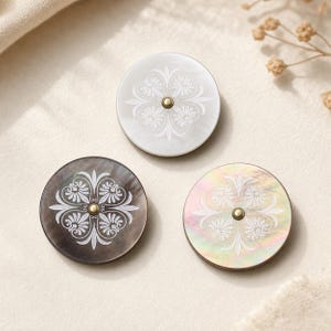 May include: Three circular buttons made of iridescent shell material, each featuring a central brass-toned stud and a white engraved floral pattern. One button is white, one is dark gray, and one displays a multi-colored pearlescent finish.