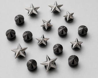May include: A collection of small body jewelry pieces consisting of silver-toned metal star-shaped studs and solid black circular studs with a pointed conical center. The items are arranged in a scattered pattern on a neutral light gray surface.