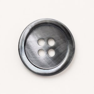 May include: A round, dark gray button with a smooth, iridescent finish and a subtle sheen. The button features a recessed center area containing four evenly spaced circular holes for attachment, surrounded by a slightly raised, smooth outer rim.