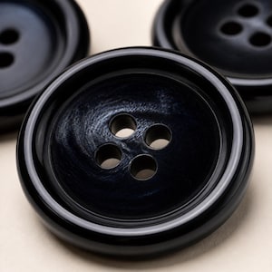 May include: A set of three round, dark navy blue buttons with a glossy finish. Each button features a raised outer rim and a recessed center with four evenly spaced holes for thread. The surface of the buttons displays a subtle, swirling texture.