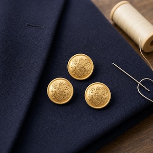 May include: Three round gold-toned metal buttons featuring an embossed heraldic crest design with a shield and intricate scrollwork. The buttons are placed on a dark navy blue fabric surface next to a wooden spool of light-colored sewing thread.