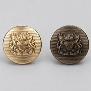 May include: Two round metal buttons featuring a raised heraldic crest design with a crown and two animals. One button has a bright gold finish, while the other has a darker, antique bronze finish. Both buttons have a fine horizontal line texture.