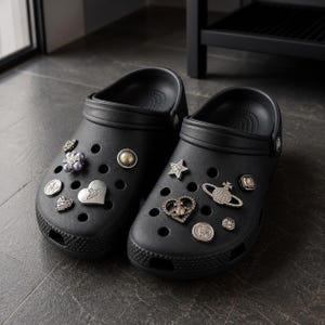 May include: A pair of black slip-on clogs featuring a series of decorative charms attached to the upper surface. The charms include metallic hearts, stars, circular studs, and a Saturn-shaped emblem with clear crystals, all set against the matte black.