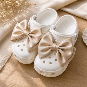May include: A pair of white slip-on clogs featuring large, champagne-colored satin bows attached to the top. The clogs have a perforated toe box, a rounded silhouette, and a textured rubber sole, providing a clean and simple design for footwear.