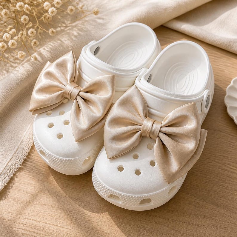 May include: A pair of white slip-on clogs featuring large, champagne-colored satin bows attached to the top. The clogs have a perforated toe box, a rounded silhouette, and a textured rubber sole, providing a clean and simple design for footwear.