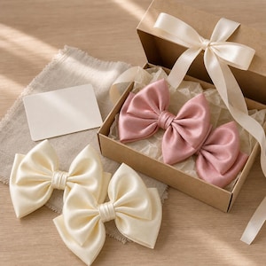 May include: A set of satin hair bows in cream and dusty rose colors. Two cream bows are placed outside a brown cardboard gift box, while two dusty rose bows are nestled inside on white tissue paper. A cream ribbon is tied around the box.