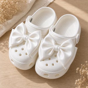 May include: A pair of white slip-on clogs featuring large, smooth satin bows attached to the top of each shoe. The clogs have a perforated toe box, a rounded silhouette, and a pivoting heel strap, all rendered in a solid, clean white finish.