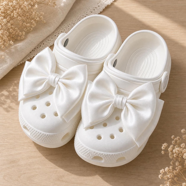 May include: A pair of white slip-on clogs featuring large, smooth satin bows attached to the top of each shoe. The clogs have a perforated toe box, a rounded silhouette, and a pivoting heel strap, all rendered in a solid, clean white finish.