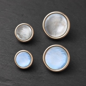May include: Four circular buttons with smooth, iridescent mother-of-pearl centers and polished gold-toned metal rims. The set includes two smaller buttons and two larger buttons, each featuring a distinct pearlescent sheen and reflective surface.