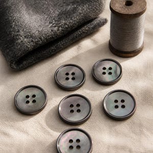 Peut inclure: Six boutons ronds gris foncé avec un léger éclat irisé, chacun doté de quatre trous centraux. Une bobine en bois entourée de fil gris et un morceau de tissu texturé gris foncé plié sont placés à proximité des boutons.