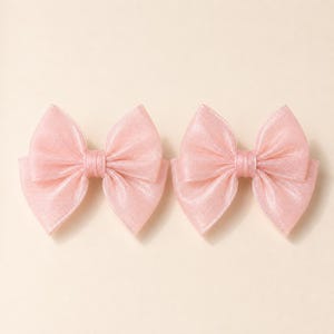 May include: Two matching hair bows in a soft, pale pink color. Each bow is constructed from a translucent, shimmering fabric with a fine, sheer texture. The bows feature a classic multi-layered design with a central knot and wide, rounded loops.