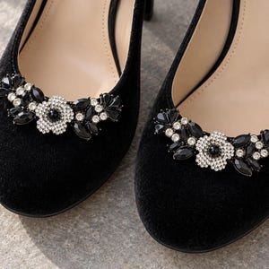 May include: A pair of black suede pumps featuring a rounded toe and a decorative embellishment on the front. The ornament consists of a central flower design surrounded by black marquise-cut crystals and small clear round rhinestones.