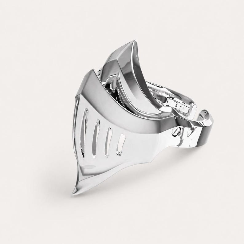 May include: A polished silver-toned ring shaped like a knight's helmet visor. The metal surface is reflective and smooth, featuring a series of vertical cutouts along the front section that mimic the appearance of a medieval armor faceplate.