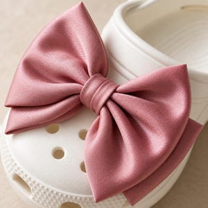 May include: A white foam clog shoe featuring a large, dusty rose-colored satin bow attached to the top. The bow is constructed from smooth, folded fabric with a central knot, while the shoe has a perforated toe box and a textured, molded surface.