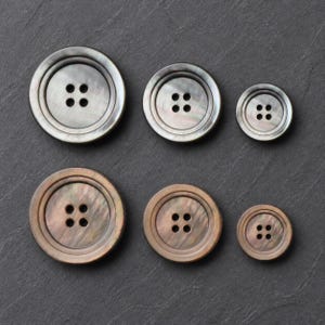 May include: A collection of six round, four-hole buttons arranged in two rows of three, varying in size from large to small. The top row features grey iridescent buttons, while the bottom row displays brown iridescent buttons with a smooth finish.