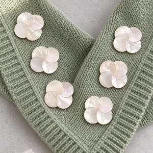 May include: Five flower-shaped buttons made of iridescent white mother-of-pearl shell, each featuring four overlapping circular petals. These decorative fasteners are arranged on a sage green knitted fabric with a ribbed border texture.