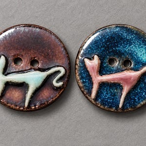 May include: Two round ceramic buttons featuring a raised cat silhouette. The left button has a glossy brown finish with a pale mint cat, while the right button has a deep blue speckled finish with a light pink cat. Each has two central holes.