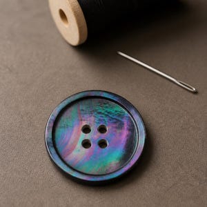 Puede incluir: Un botón redondo de cuatro agujeros con una superficie oscura e iridiscente que presenta tonos arremolinados de morado, azul y verde. Junto al botón hay una aguja de coser plateada y un carrete de madera con hilo negro.
