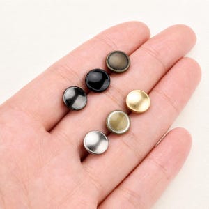 Può includere: Una collezione di sei piccoli bottoni metallici circolari disposti su due file. I bottoni presentano varie finiture metalliche, tra cui argento lucido, oro opaco, bronzo scuro e nero lucido, ciascuno con una superficie liscia e bombata.