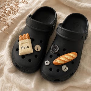 Könnte beinhalten: Ein Paar schwarze Clogs mit dekorativen Ansteckern. Ein Clog hat einen beigen Taschen-Anstecker mit dem Text "Pain" und einen kleinen runden Anstecker. Der andere Clog zeigt einen Baguette-Anstecker und zwei runde knopfartige Anstecker mit Löchern.