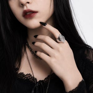 May include: A silver-toned ring featuring a stylized helmet design with vertical cutouts. The ring has a polished metallic finish and a pointed top section, worn on the finger. The design resembles a knight's visor, crafted from solid metal.