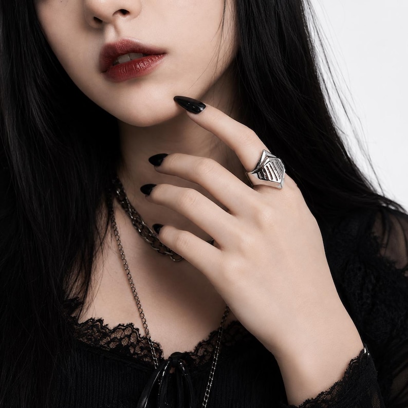 May include: A silver-toned ring featuring a stylized helmet design with vertical cutouts. The ring has a polished metallic finish and a pointed top section, worn on the finger. The design resembles a knight's visor, crafted from solid metal.