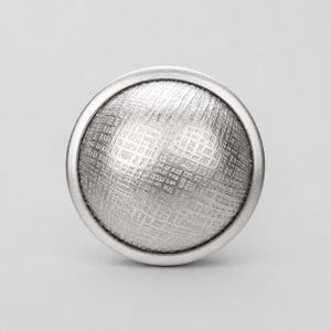 May include: A circular silver-toned cabinet knob featuring a domed front surface with a textured, cross-hatched grid pattern. The knob has a smooth, polished outer rim that frames the detailed, etched centre, creating a clean and uniform appearance.