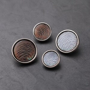May include: Four circular buttons with metallic silver rims and textured, swirling patterns. Two buttons feature a dark brown finish, while the other two display a shimmering, iridescent light gray finish, all arranged in a cluster on a dark surface.