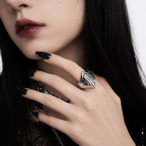 May include: A silver-toned metal ring shaped like a medieval knight's helmet visor. The ring features a pointed top and vertical slits across the front surface, with a polished, reflective finish that catches the light on the finger.