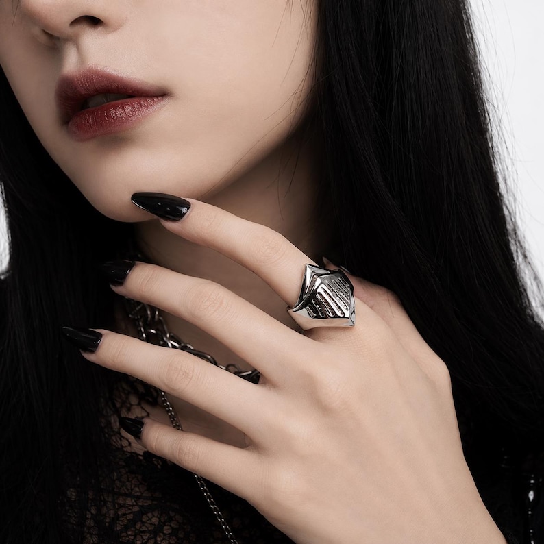 May include: A silver-toned metal ring shaped like a medieval knight's helmet visor. The ring features a pointed top and vertical slits across the front surface, with a polished, reflective finish that catches the light on the finger.