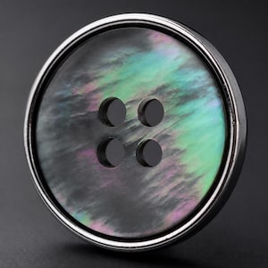 Puede incluir: Un botón redondo de cuatro agujeros con un centro de nácar oscuro e iridiscente con reflejos verdes y morados, encerrado en un borde de metal plateado liso y pulido que proporciona un marco limpio y reflectante alrededor de la concha.