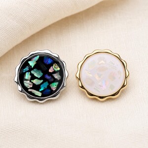 May include: Two circular buttons with scalloped edges. The left button has a silver-tone rim and a black inlay filled with iridescent, multi-colored shell fragments. The right button has a gold-tone rim and a white inlay with shell pieces.