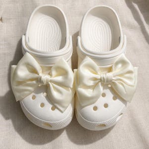 May include: A pair of off-white slip-on clogs featuring large, decorative satin bows attached to the top of each shoe. The clogs have a rounded toe, a series of ventilation holes across the vamp, and a molded footbed with a textured surface.