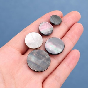 Puede incluir: Cinco botones circulares de tamaños variados con un acabado liso e iridiscente. Los botones muestran una gama de colores que incluye tonos gris oscuro, carbón y rosa suave, con un brillo nacarado reflectante en sus superficies planas.