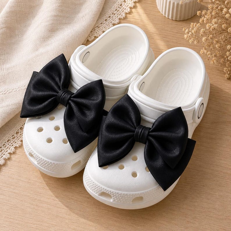 May include: A pair of white slip-on clogs featuring large, black satin-like bows attached to the top of each shoe. The clogs have a perforated toe box, a rounded silhouette, and a pivoting heel strap, showcasing a clean, solid white base.