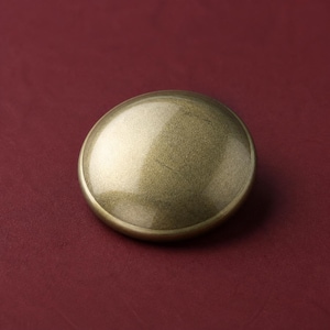 May include: A circular, dome-shaped button with a smooth, metallic finish in a muted antique gold or brass tone. The surface has a subtle, reflective sheen and a uniform, slightly weathered texture across its convex, rounded top profile.
