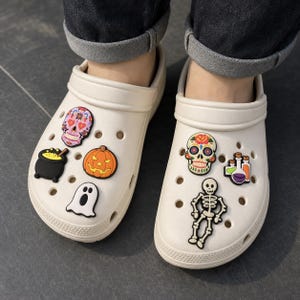 May include: A pair of off-white slip-on clogs decorated with various Halloween-themed charms. The charms include a colorful sugar skull, a black cauldron with yellow liquid, an orange jack-o'-lantern, a white ghost, a skeleton, and potion bottles.