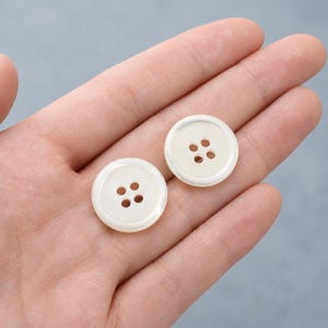 May include: Two round, off-white buttons with a smooth, pearlescent finish. Each button features a slightly raised outer rim and four small, evenly spaced holes in the center for attachment. The buttons have a glossy, reflective surface texture.