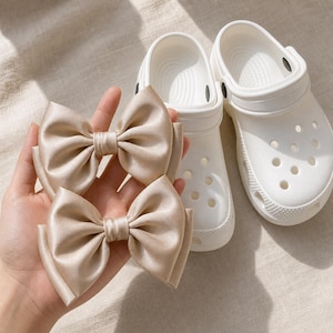 May include: Two champagne-colored satin bows with a smooth, lustrous finish, designed as decorative shoe charms, positioned next to a pair of white rubber clogs featuring a perforated upper and a pivoting heel strap for a secure fit.
