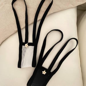 Könnte beinhalten: Zwei Tanga-Unterwäschestücke mit schwarzen elastischen Bändern und Einfassungen. Ein Modell hat ein Vorderteil aus transparentem weißem Netzstoff, das andere ein blickdichtes schwarzes Vorderteil. Jedes Stück ist mit einer kleinen weißen Blütenapplikation verziert.