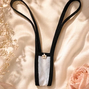 Könnte beinhalten: Schwarz-weiße Dessous mit einem Blumenschmuck. Der String-Tanga hat eine schwarze Einfassung und Träger mit einem weißen Mesh-Einsatz in der Mitte. Ein kleiner, goldfarbener Blumenschmuck hängt vorne.