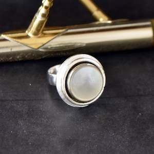 May include: A silver ring with a white mother-of-pearl stone set in a circular bezel. The ring has a textured band.