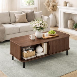 May include: A dark brown wooden coffee table with a slatted front and a lower shelf. The table has black legs and is topped with a white vase, a mug, and a tray of fruit. The table is in a living room setting.
